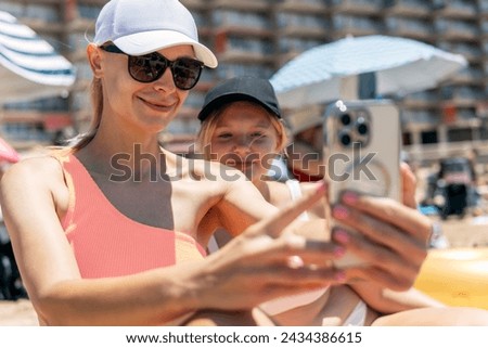 A mother and teenage daughter in swimsuits take a selfie against the backdrop of a summer beach on their phone, capturing cherished moments together.