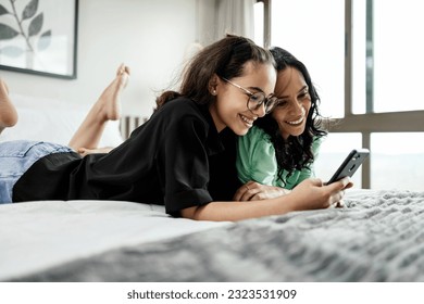 Mother and teenage daughter using smartphone together lying in bed at home. Latina mother and daughter looking at phone watching social media videos while relaxing in bedroom. family and technology - Powered by Shutterstock