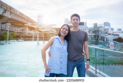 Mother And Teenage Son, Tall, Well-proportioned, Good-looking, Healthy, Standing Around The Waist, Embracing Each Other, Smiling At The Camera. There Was An Orange Light Shining From Behind.