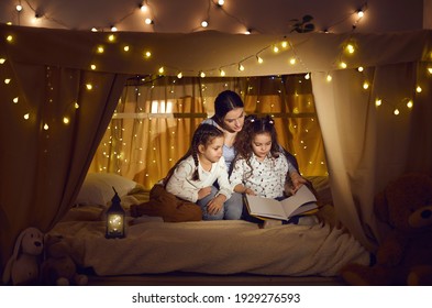 Mother Telling Bedtime Story To Children. Mommy And Little Daughters Sitting In Magic Blanket Fort In Cozy Dark Playroom, Reading Book Of Good Fairy Tales And Enjoying Quiet Family Evening At Home