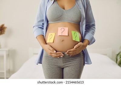 Mother thinking of name for future baby. Woman trying to guess gender of unborn child. Pregnant lady with question mark sticky notes on big bare belly, cropped shot. Name choice, gender reveal concept