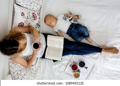 Mother And Toddler Baby Boy, Lying In Bed, Baby Sleeping, Mom Reading A Book And Eating Fruits And Ice Cream, High Angle Shot, From Above. Happy Family Concept
