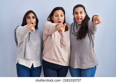 Mother And Two Daughters Standing Over Blue Background Pointing With Finger Surprised Ahead, Open Mouth Amazed Expression, Something On The Front 