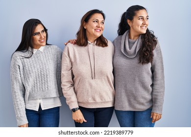 Mother And Two Daughters Standing Over Blue Background Looking Away To Side With Smile On Face, Natural Expression. Laughing Confident. 