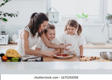 Mother And Two Smiling Daughters Making Pancakes In A White Kitchen, Having Fun, Dressed In White, Together Family Time.