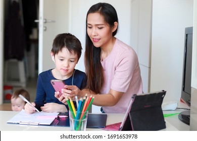 Mother using smartphone and tablet doing homework with her kid self isolation during coronavirus, covid-19 lock down quarantine, social distancing, online E-learning education. Home schooling