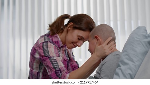 Mother visiting teen son with cancer in hospital ward. Side view of bald kid resting in hospital bed and hugging mother visitor. Healthcare and medicine concept