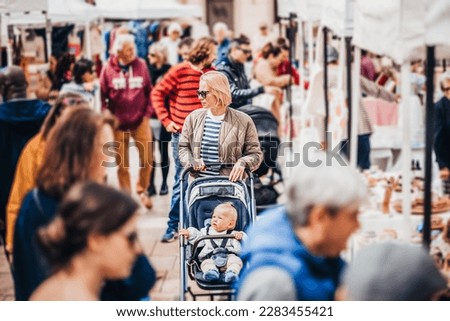 Mother waling and pushing his infant baby boy child in stroller in crowd of unrecognizable people wisiting sunday flea market in Malaga, Spain
