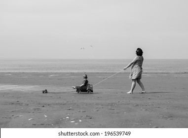 a mother walking with her son on the beach in black and white. She is pulling a car where the boy is in. The boy is pulling on his own little car. Two seagulls are flying in the sky.