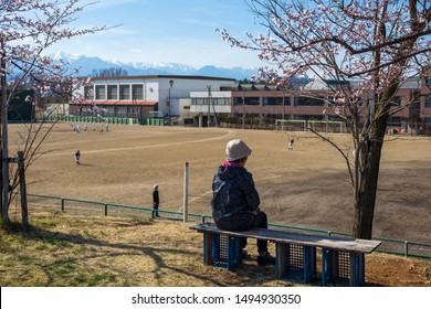 Mother Watching Her Son To Play Baseball In Team With Cherry Blossom Or Sakura Flowers And Central Alps Mountain, Matsumoto, Japan. Outdoor Sport Activity Of Highschool Students.