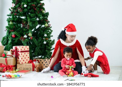 Mother Wear Santa Hat And Black Baby Son With Pacifier, Daughter Hugging And Having Fun In Front Of The Christmas Tree And Presents. New Year's And Christmas Eve. Cozy Holiday With Mixed Race Family.