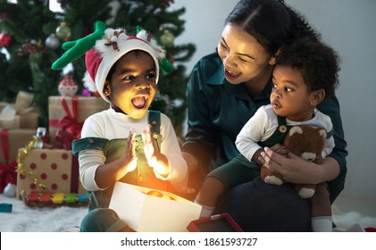 Mother Wear Santa Hat And Black Baby Son With Pacifier, Daughter Hugging And Having Fun In Front Of The Christmas Tree And Presents. New Year's And Christmas Eve. Cozy Holiday With Mixed Race Family.