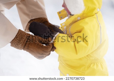 A mother woman puts a mitten on the hand of a toddler baby boy. The parent helps the child to put on a glove, warm winter clothes. Kid age one year eight months