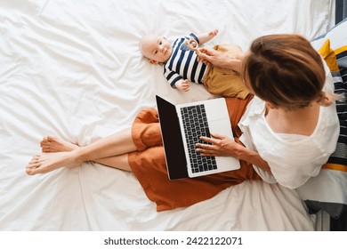 Mother working at laptop playing with baby in bed at home. Modern woman balances between work and baby. Child playing with toy on bed while mother using laptop: surfing internet, doing online shopping - Powered by Shutterstock