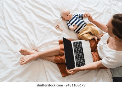 Mother working at laptop playing with baby in bed at home. Modern woman balances between work and baby. Child playing with toy on bed while mother using laptop: surfing internet, doing online shopping - Powered by Shutterstock