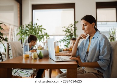 Mother working on laptop at kitchen table while children have breakfast. Remote work and home office for mothers with children.