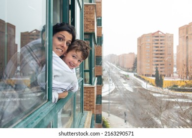 Mother Wrapped In A Warm White Blanket Hugging Her Child And Looking Out The Window During A Cold Winter Snow Storm.