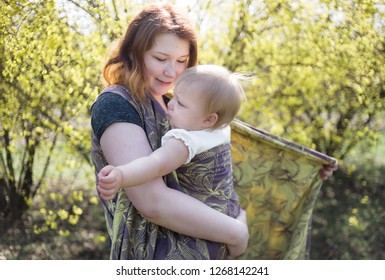 Mother Wrapping Her Baby Daughter In A Sling For Babywearing. High Key, Lifestyle Image Shot On Location In A Park On A Sunny Spring Day. 