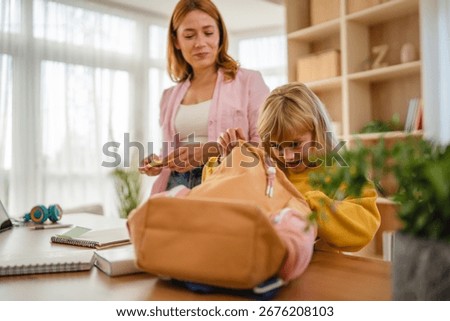 Mother writing a note while helping her surprised daughter pack her school backpack at home, creating a warm morning routine filled with love and support for learning together