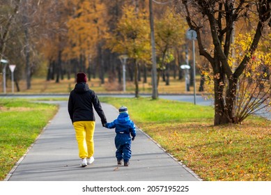 A Mother In Yellow Trousers With A Small Cute Child Are Walking In The Autumn Park. Walking And Clean Air Are Good For Your Health. 