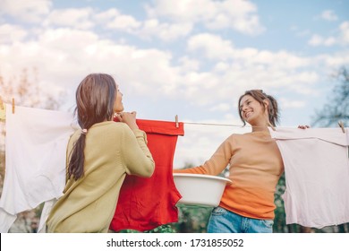 Mother And Young Daughter Hang Clothes Outdoors To Dry. Smiling Daughter Helps Mom With Laundry. Cheerful Mother And Daughter In Conversations. Woman Clothes Are Drying. Celebration. Happy Day. Life.