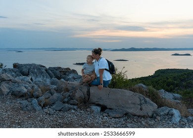 Mother and young son enjoying sunset on rocky hilltop with scenic sea and island view. Concept of family bonding, outdoor adventure, and nature appreciation. High quality photo - Powered by Shutterstock