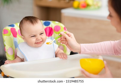 Mothercare. Cheerful Baby Eating In Kitchen With Mother Help, Sitting In High Chair, Empty Space