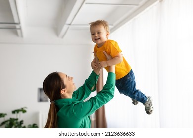 Motherhood Concept. Playful Lady Holding Her Smiling Child Boy, Lifting Son High Up In The Air, Woman Playing With Baby And Having Fun Together At Home, Free Space