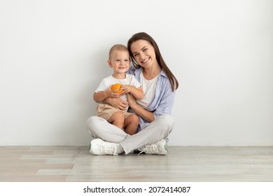 Motherhood Concept. Portrait Of Beautiful Young Woman Sitting On Floor And Embracing Her Cute Infant Son, Loving Mom Enjoying Time With Her Baby, Looking And Smiling At Camera, Copy Space