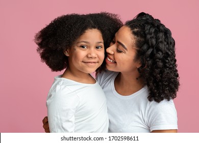 Mother's care. Smiling african woman tenderly cuddling her cute little daughter, pink background with free space