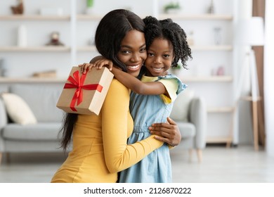 Mother's Day Concept. Adorable Female Child Holding Gift And Embracing Mom, Portrait Of Happy African American Mommy And Her Cute Little Daugher Bonding Together At Home, Copy Space