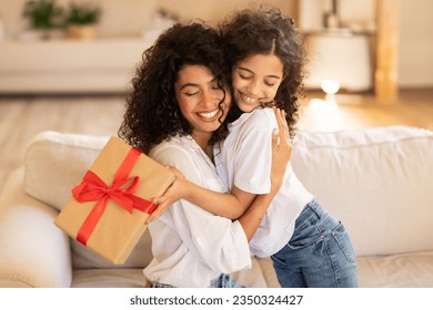 Mother's day concept. Adorable latin girl giving gift and embracing mom, mommy and daughter bonding together at home, sitting on sofa - Powered by Shutterstock