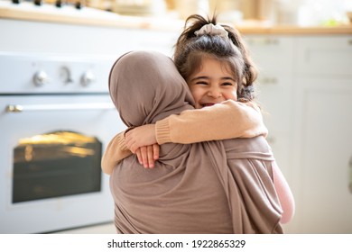 Mother's Day Concept. Cheerful Little Girl Hugging Tight Her Muslim Mom In Hijab, Islamic Woman Embracing Smiling Female Child, Happy Family Bonding Together In Kitchen At Home, Copy Space