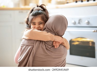 Mother's Day Concept. Cheerful Little Girl Hugging Tight Her Muslim Mom In Hijab, Islamic Woman Embracing Smiling Female Child, Happy Family Bonding Together In Kitchen At Home, Copy Space