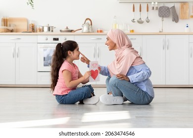 Mother's Day Concept. Portrait Of Loving Little Girl Presenting Handmade Card To Her Muslim Mom In Hijab, Happy Islamic Mommy With Cute Female Child Sitting On Floor In Kitchen, Bonding At Home