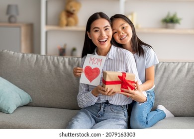Mother's Day gift. Happy asian girl greeting young surprised mom, giving her handmade card and wrapped gift box, sitting on sofa at home interior and smiling at camera