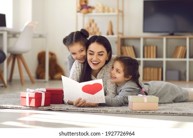 Mother's Day. Twin sisters hug and greet their mother by giving her a handmade card and gifts. Excited happy mother lying on the floor with children and reading greetings. Concept of family and love.