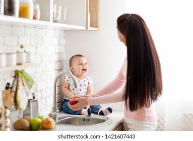 Mother's little helper. Adorable baby sitting on table in kitchen while mom washing apple for snack, empty space