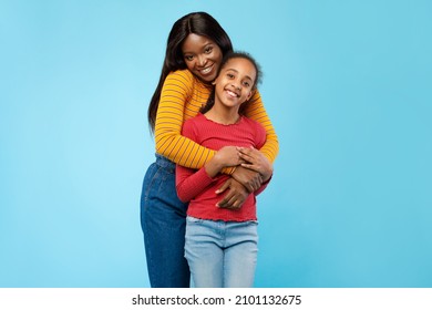 Mother's Love Concept. Portrait of happy young African American mom cuddling her cute tween daughter from the back, standing posing isolated over blue studio background with copy free space