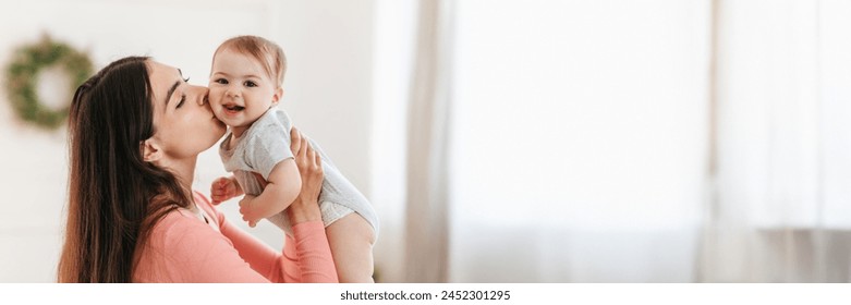 Mother's love. Happy young mother holding her child in hands and kissing in cheek while playing and enjoying time with kid at home. Happy maternity leave concept, panorama with copy space - Powered by Shutterstock