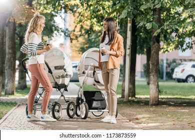mothers standing with coffee to go near baby strollers in park 
