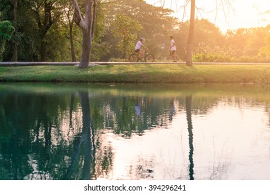 Motion Blured Of People Are Running And Cycling In Urban Park With Water Reflection. 