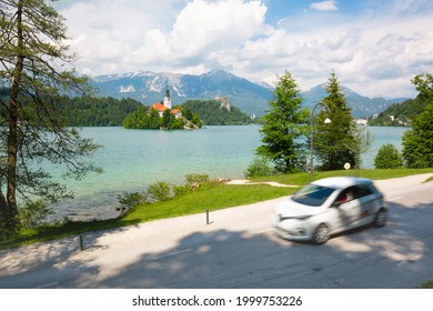 Motion Blured White Electric Car In Front Of Bled Lake, Country Of Slovenia. Travel, Tourism, Explore, Rent A Car Concept.