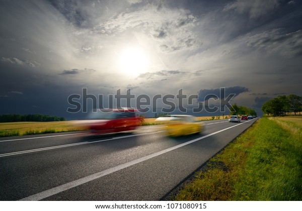 Motion\
blurred cars driving on a road in a rural landscape under the sun\
shining through the clouds before the\
storm