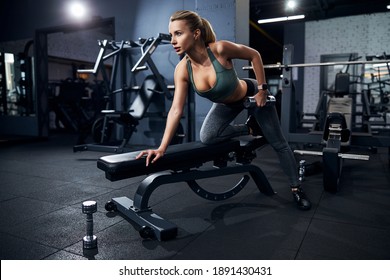 Motivated athletic citizen supporting herself with a right hand on a bench and doing the dumbbell lifts - Powered by Shutterstock