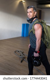 Motivated Boot Camp Instructor Stands With Gym Equipment In Gym Hall. Carry Training Mask, Dumbbells And Sandbag, Rope On Wooden Floor. Instructor Smiling To Camera. Portrait For Gym Concept.