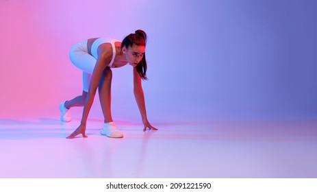 Motivated Female Runner Standing In Crouch Start Position Ready For Race Over Pink And Blue Neon Background. Studio Shot Of Sporty Woman In Fitwear Preparing For Running Posing In Studio