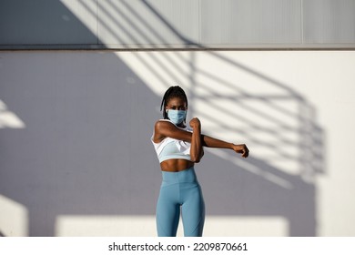 Motivated Female Urban Athlete With Face Mask Training Outside Under Coronavirus Pandemic. Shoulder Stretching Warm Up Exercise Before Running.