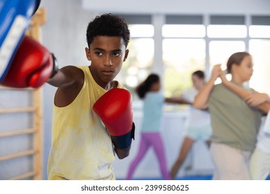 Motivated focused african american boy wearing boxing gloves working out with coach during group self-defense course, practicing punches on mitts.. - Powered by Shutterstock