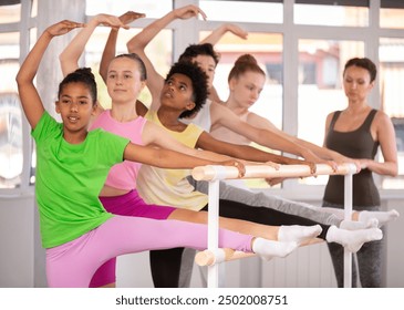 Motivated interested dark-skinned girl with group of teenagers stretching at ballet barre during beginner dance class, guided by female instructor, in light-filled choreography studio - Powered by Shutterstock